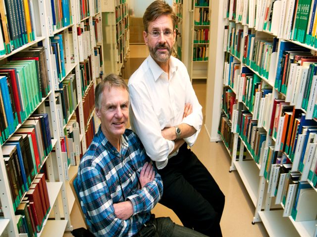To menn sitter i et bibliotek og smiler.