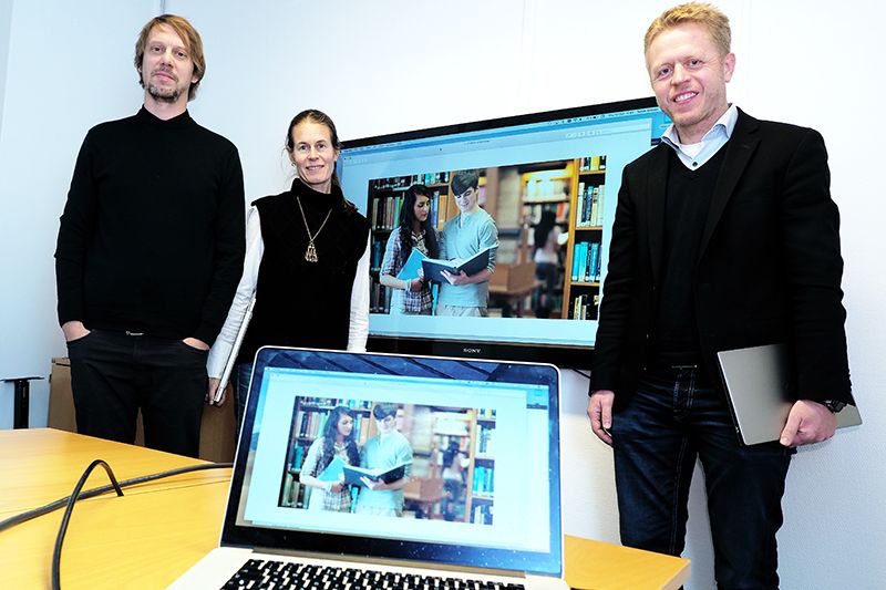 To menn med ei kvinne midten står framfor ein storskjerm med ein PC-skjerm i forgrunnen
