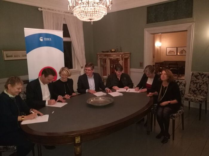 Sju personar sit rundt eit bord og underskriv ein samarbeidsavtale.