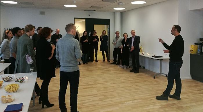 Ein mann står og snakkar medan han dirigerer med hendene medan fleire personar står i ring i møtelokalet.