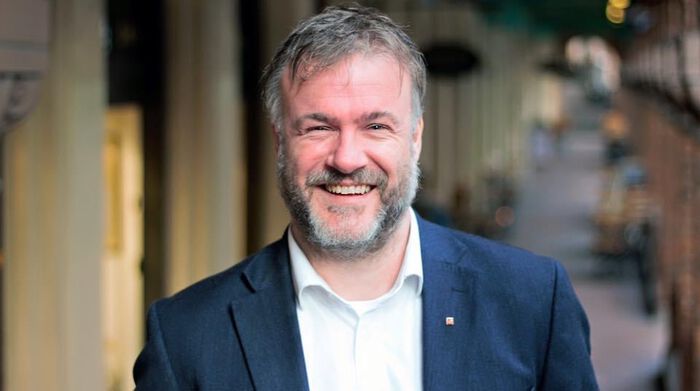 En smilende mann med skjegg står og har en gate i bakgrunnen