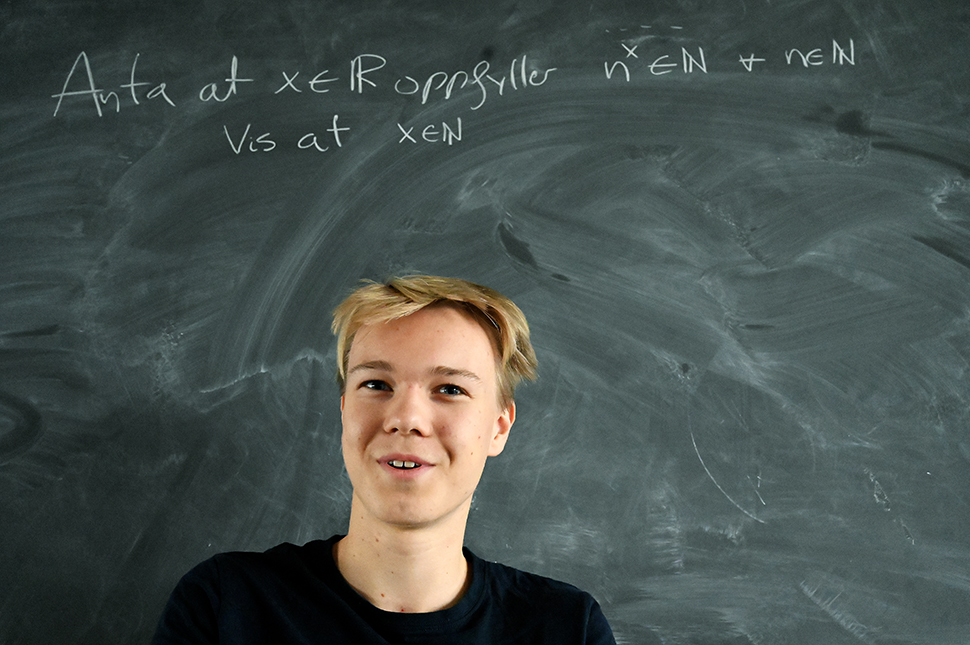 Ein ung gut ser mot kameraet med ei tavle med ei skriftleg matematikkoppgåve i bakgrunnen,