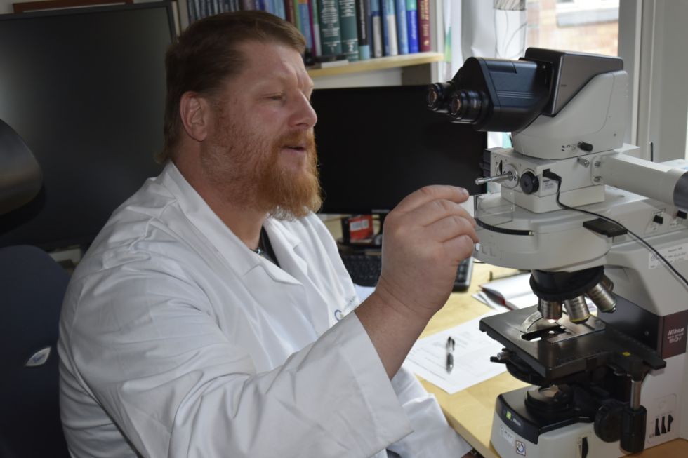 Ein mann i kvit frakk sit og kikar i  eit mikroskop