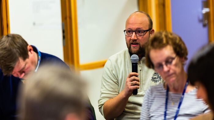 En mann med briller og mikrofon tar ordet på et fellesmøte