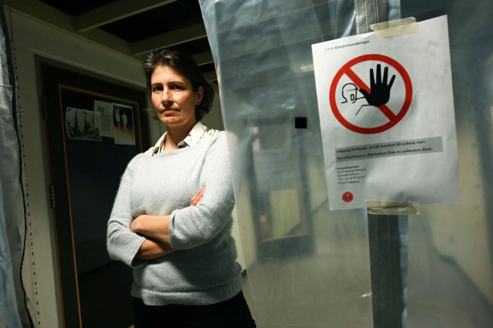 Ei kvinne står med armane i kryss ved sidan av eit varselskilt om asbest